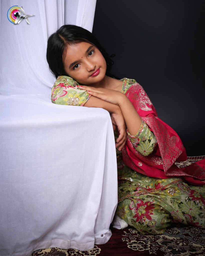 A stylish girl in a floral printed green kurta-palazzo set with a contrasting red dupatta poses in a studio setting with curtains and decor beside her