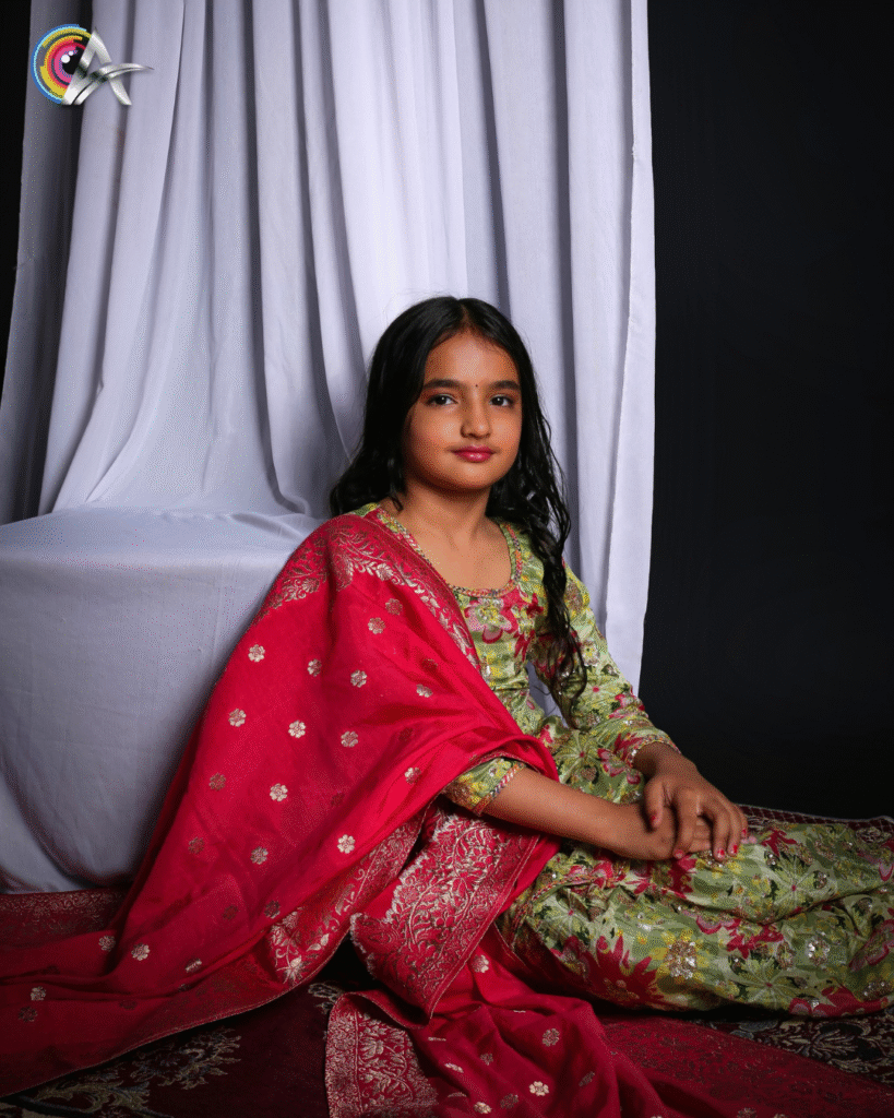 Elegant young girl showcasing Indian ethnic wear with a floral print and rich red dupatta, striking a graceful pose in a studio photoshoot with cultural elements.