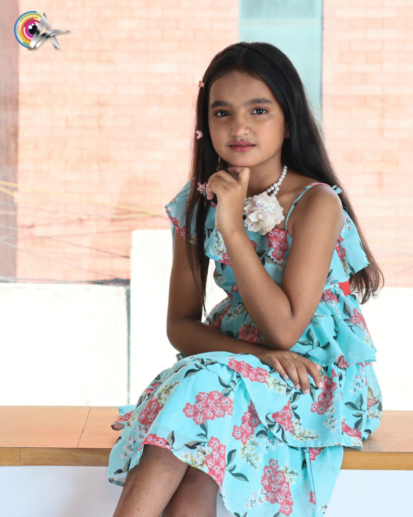 Young girl posing indoors, wearing a flowery blue dress with pink accents.