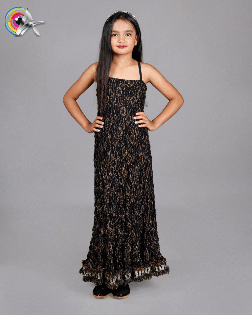 A young girl with long straight hair wearing a black and gold sleeveless floor-length gown with textured patterns. She is standing sideways with one hand touching her face and the other folded, looking confident. She is also wearing a headband and black shoes with golden soles.