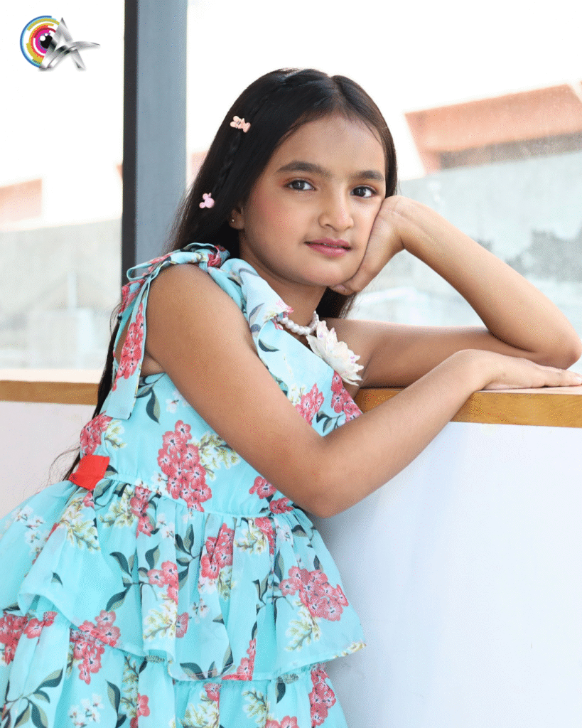 young girl with long black hair, wearing butterfly hair clips and a blue floral dress with pink and white flowers, poses by resting her elbow on a ledge and her chin on her hand, smiling softly at the camera.