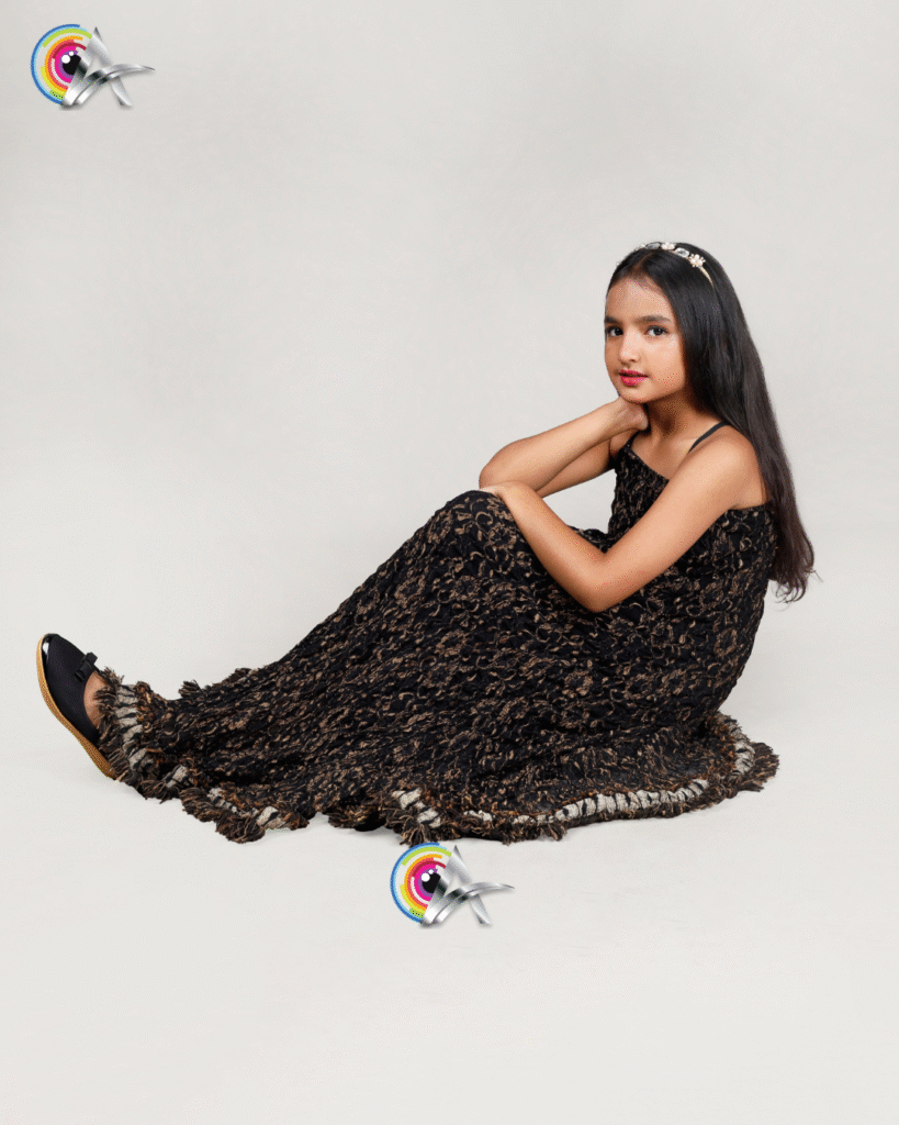 Elegant young girl posing in a sleeveless black and gold textured floor-length gown with a flared hem, styled with a jeweled headband and black shoes, captured in a professional kids’ fashion photoshoot.