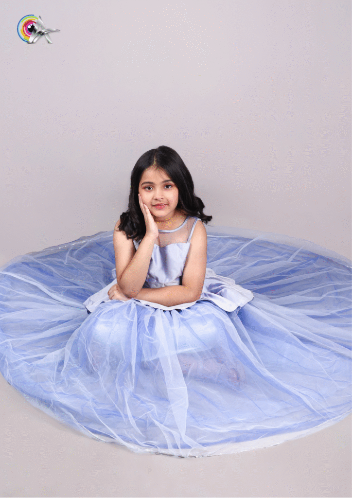 Young child model in a stylish blue dress with a peplum top and long net skirt on a plain background.